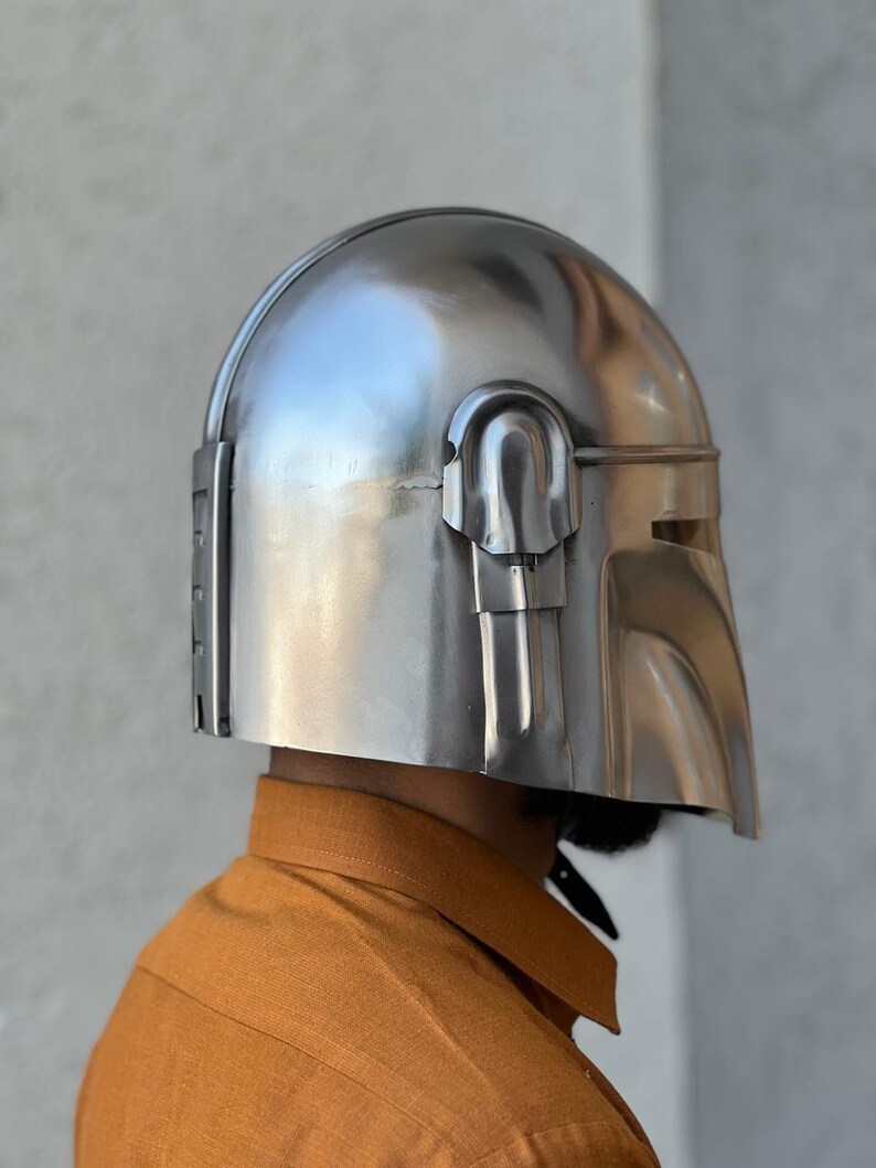 May include: A silver helmet with a unique visor and rounded crown. The metallic helmet has a reflective surface. The design includes vertical lines on the side and a curved visor opening. The helmet is likely made of metal.