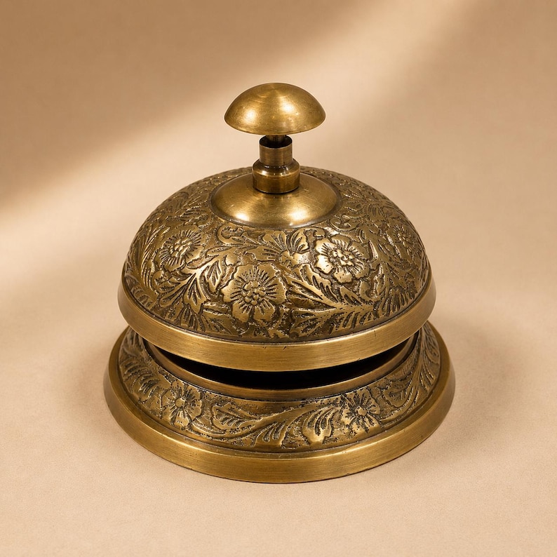 Peut inclure: Une cloche de bureau en laiton de style vintage orn&eacute;e d'un motif floral en relief complexe sur son d&ocirc;me et sa base. La cloche pr&eacute;sente une finition m&eacute;tallique polie avec un bouton-poussoir arrondi, con&ccedil;ue pour une utilisation manuelle.