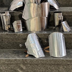 May include: A collection of polished silver metal armor pieces, including a chest plate, shoulder guards, and arm and leg protection. The armor is displayed on a set of gray steps, showcasing its reflective surface and intricate design.