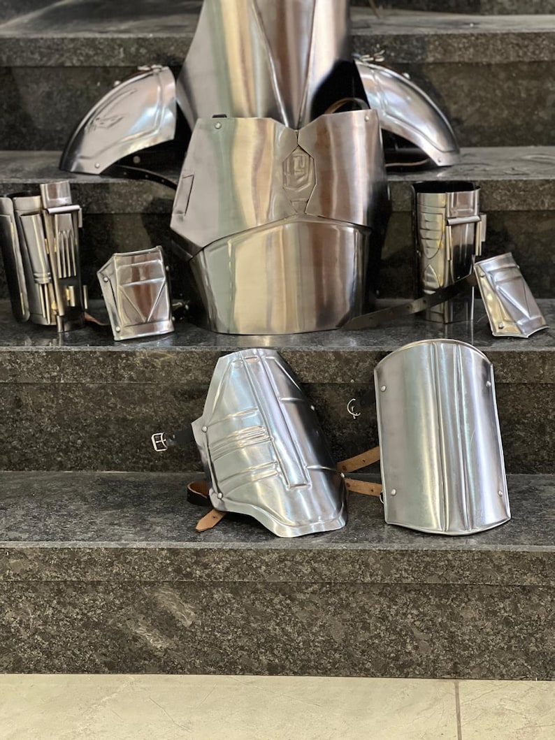 May include: A collection of polished silver metal armor pieces, including a chest plate, shoulder guards, and arm and leg protection. The armor is displayed on a set of gray steps, showcasing its reflective surface and intricate design.