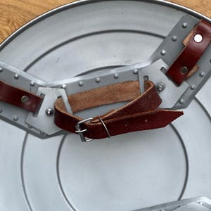 K&ouml;nnte beinhalten: Ein silberfarbener, kreisf&ouml;rmiger Schild mit genieteten grauen Metallakzenten und braunen Lederriemen. Der Schild hat ein zentrales Design mit konzentrischen Kreisen und ist mit einem Griff versehen.