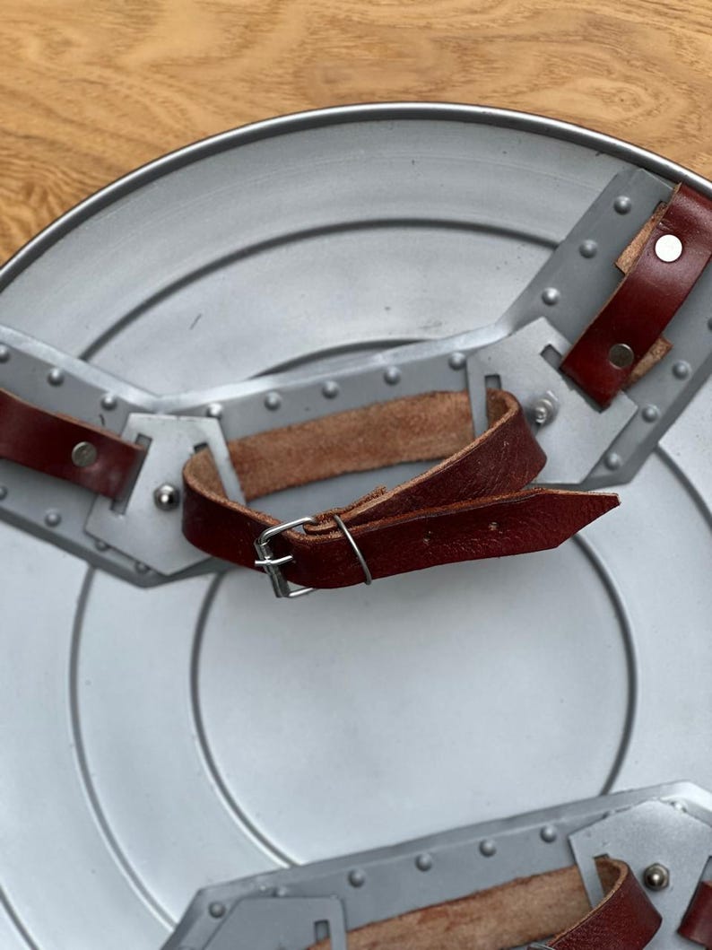 K&ouml;nnte beinhalten: Ein silberfarbener, kreisf&ouml;rmiger Schild mit genieteten grauen Metallakzenten und braunen Lederriemen. Der Schild hat ein zentrales Design mit konzentrischen Kreisen und ist mit einem Griff versehen.
