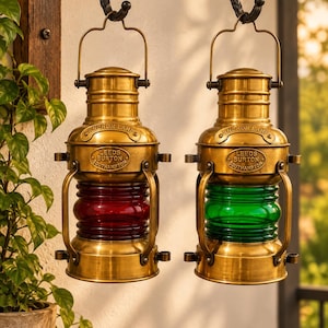 May include: Two brass anchor lamps hanging from a black metal wall bracket. Each lamp features a cylindrical body with a central glass section, one red and one green. The metal plates on the lamps are embossed with the text ANCHOR LAMP LEEDS BURTON SOUTHAMPTON.