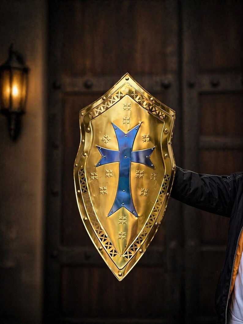 Op de afbeelding: Een goudkleurig schild met een blauw kruisontwerp. Het schild heeft een decoratieve rand en stervormige accenten. Het schild wordt vastgehouden door een persoon met een zwarte jas.