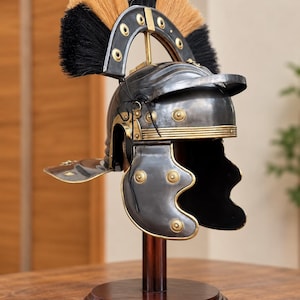 Op de afbeelding: Een metalen helm in Romeinse stijl met een donkergrijze afwerking en messingkleurige accenten, waaronder ronde klinknagels en randen. De helm is voorzien van een grote, gebogen kam gemaakt van stijve zwarte en beige borstelharen op een houten standaard.