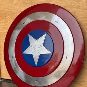 K&ouml;nnte beinhalten: Ein runder Schild mit einem roten Au&szlig;enring, einem silbernen Innenring und einer blauen Mitte. In der Mitte befindet sich ein wei&szlig;er f&uuml;nfzackiger Stern. Der Schild ist eine Nachbildung von Captain Americas Schild.