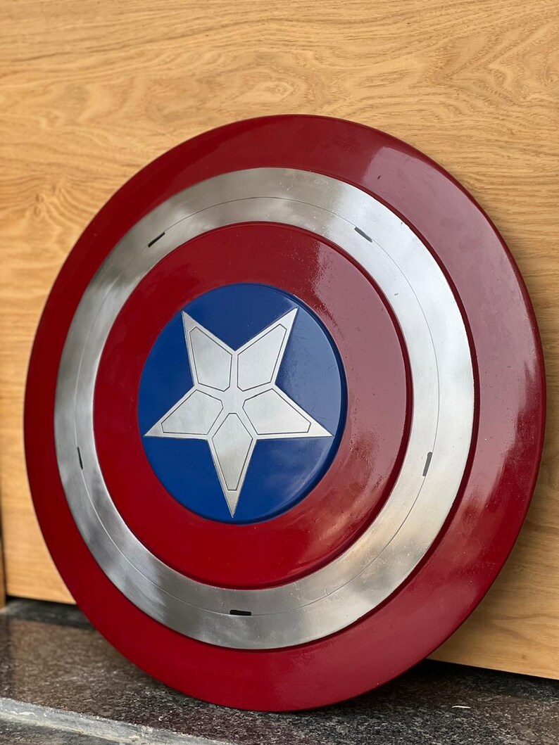 K&ouml;nnte beinhalten: Ein runder Schild mit einem roten Au&szlig;enring, einem silbernen Innenring und einer blauen Mitte. In der Mitte befindet sich ein wei&szlig;er f&uuml;nfzackiger Stern. Der Schild ist eine Nachbildung von Captain Americas Schild.