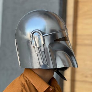 May include: A polished silver helmet with a visor and a curved design. The helmet has a smooth, reflective surface, likely made of metal. It is a costume piece, possibly for cosplay or a themed event.