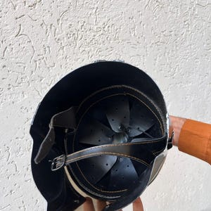 May include: A black and silver helmet with a leather strap and buckle. The interior features black leather padding and a riveted design. The helmet is held by a person.