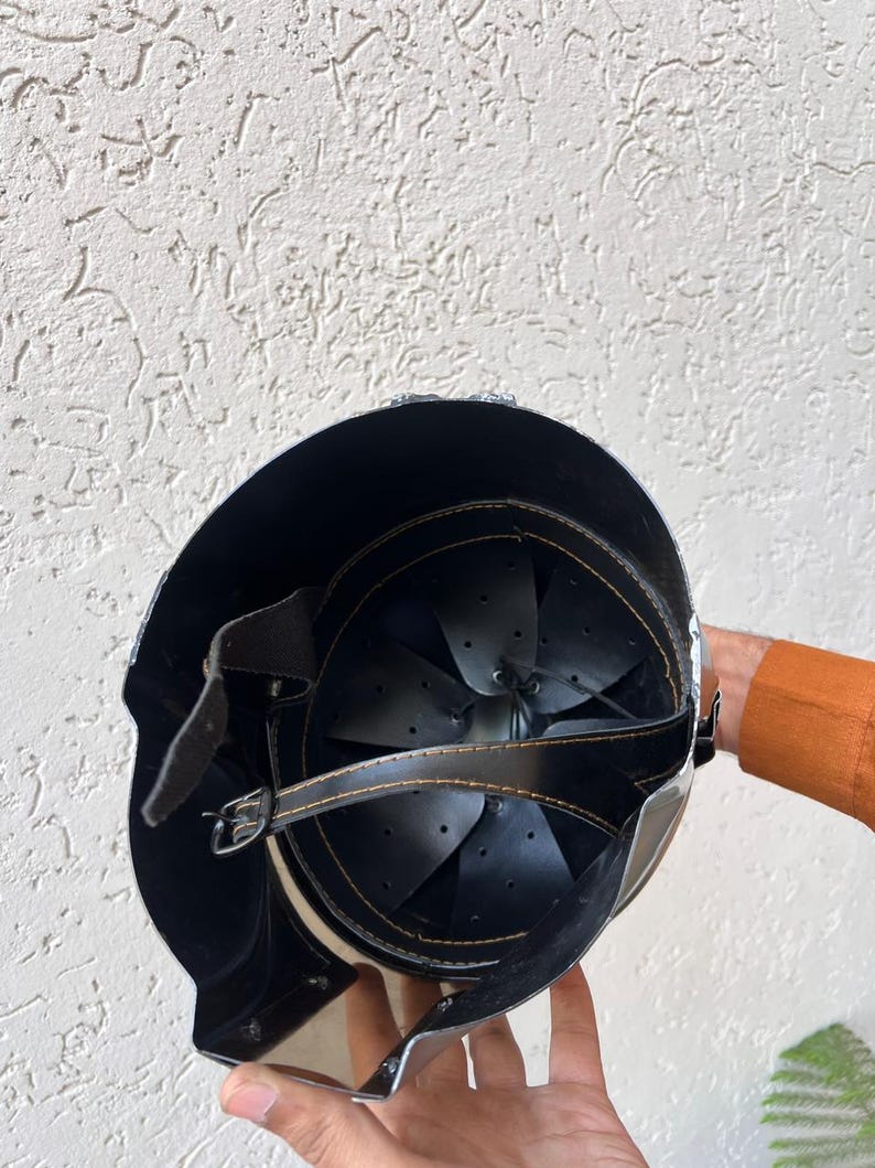 May include: A black and silver helmet with a leather strap and buckle. The interior features black leather padding and a riveted design. The helmet is held by a person.