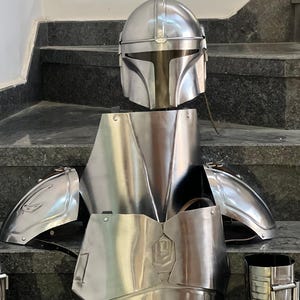 May include: A complete set of shiny, silver-colored Mandalorian armor, including a helmet, chest plate, shoulder pads, and leg guards. The armor is displayed on a dark surface, showcasing its detailed design and metallic finish.