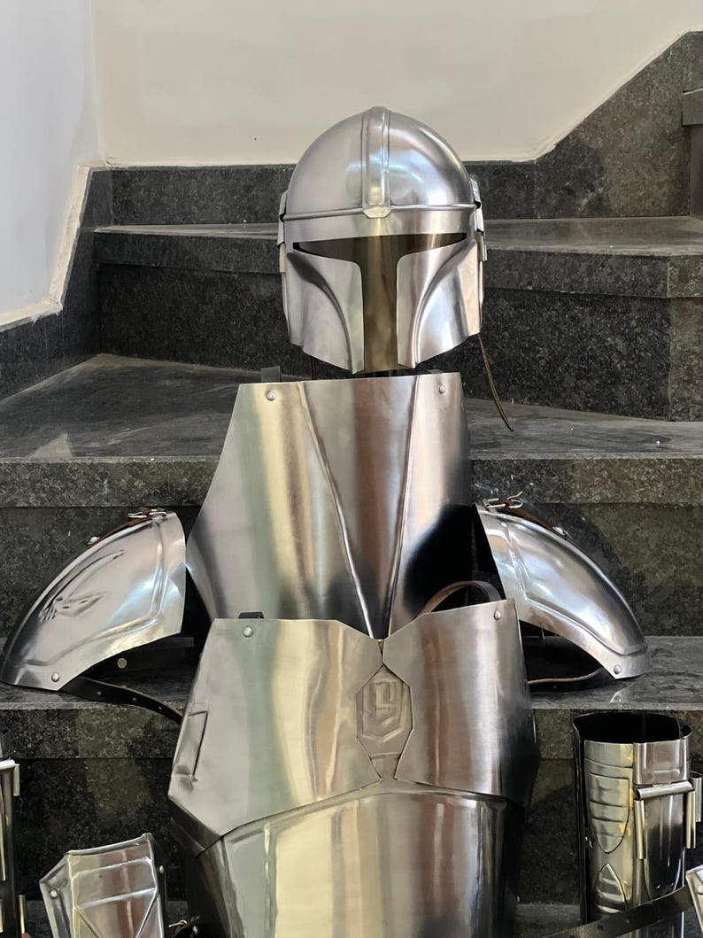 May include: A complete set of shiny, silver-colored Mandalorian armor, including a helmet, chest plate, shoulder pads, and leg guards. The armor is displayed on a dark surface, showcasing its detailed design and metallic finish.
