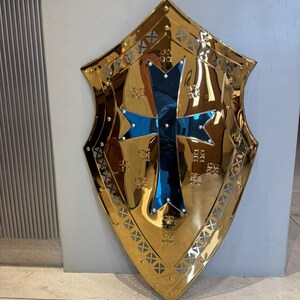 Op de afbeelding: Een goudkleurig schild met een blauw kruis in het midden. Het schild heeft een decoratieve rand en is waarschijnlijk van metaal. Het schild is een decoratief item.