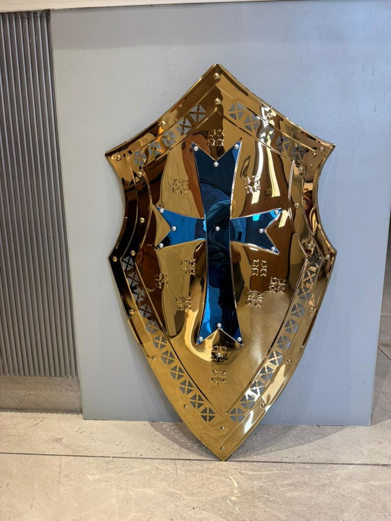 Op de afbeelding: Een goudkleurig schild met een blauw kruis in het midden. Het schild heeft een decoratieve rand en is waarschijnlijk van metaal. Het schild is een decoratief item.