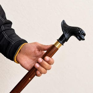 Könnte beinhalten: Ein Gehstock mit einem dunkelbraunen Holzschaft mit kleinen goldfarbenen Einlagen, einem polierten Messingring und einem schwarzen Griff in Form eines stilisierten Tierkopfes mit spitzen Ohren und ausgeprägten Gesichtszügen.