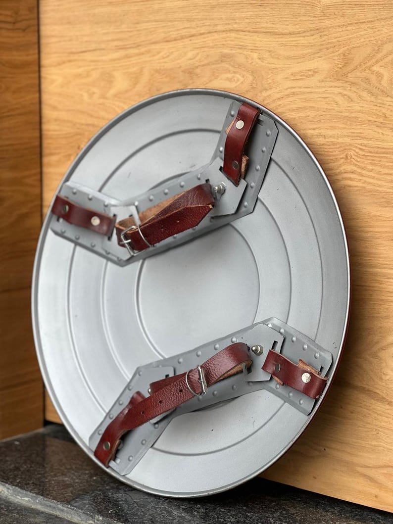 K&ouml;nnte beinhalten: Ein silberfarbener, kreisf&ouml;rmiger Schild mit konzentrischen Ringen. Es hat graue Metallhalterungen mit Nieten und braune Lederriemen. Der Schild ist eine Nachbildung eines ikonischen Accessoires eines Superhelden.