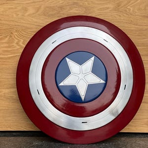 K&ouml;nnte beinhalten: Ein runder Schild mit einem roten Au&szlig;enring, einem silbernen Band und einer blauen Mitte. In der Mitte befindet sich ein wei&szlig;er f&uuml;nfzackiger Stern. Der Schild ist eine Nachbildung von Captain Americas Schild.