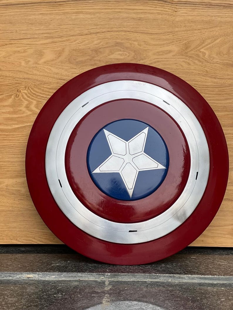 K&ouml;nnte beinhalten: Ein runder Schild mit einem roten Au&szlig;enring, einem silbernen Band und einer blauen Mitte. In der Mitte befindet sich ein wei&szlig;er f&uuml;nfzackiger Stern. Der Schild ist eine Nachbildung von Captain Americas Schild.