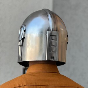 May include: A silver helmet with a rounded crown and angular side features. The helmet is made of a metallic material with a smooth, reflective surface. The person wearing the helmet is wearing an orange shirt.