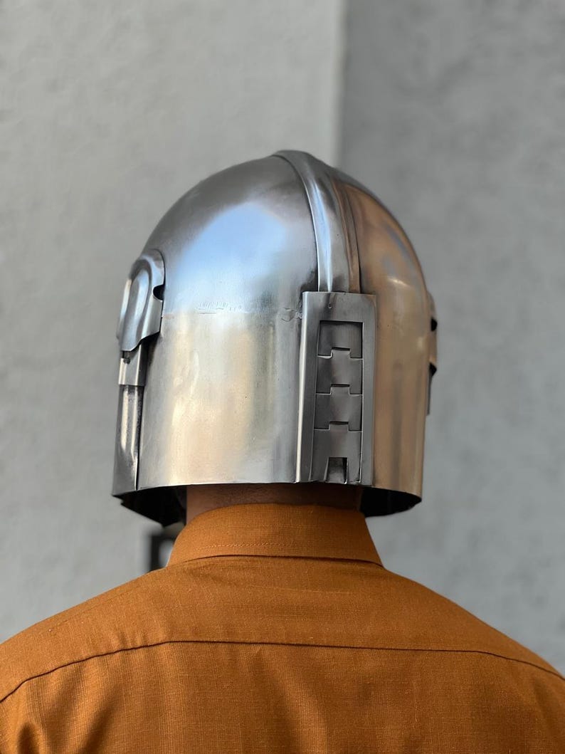 May include: A silver helmet with a rounded crown and angular side features. The helmet is made of a metallic material with a smooth, reflective surface. The person wearing the helmet is wearing an orange shirt.