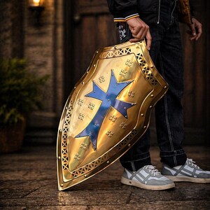 Op de afbeelding: Een goudkleurig schild met een blauw kruisontwerp. Het schild heeft een decoratieve rand en wordt door een persoon vastgehouden. Dit is een item met een middeleeuws thema.