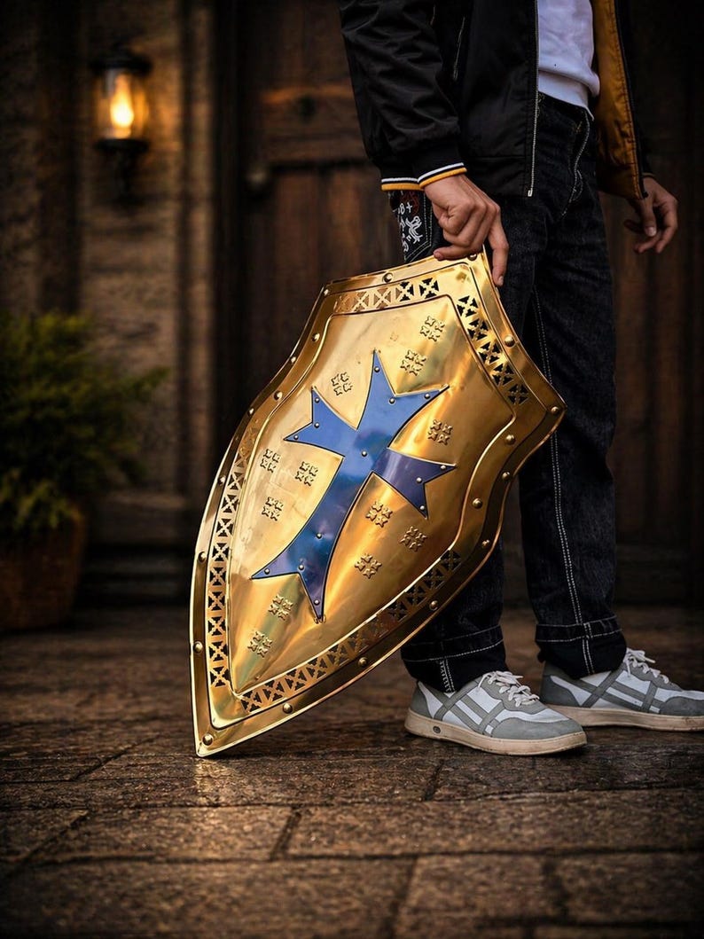 Op de afbeelding: Een goudkleurig schild met een blauw kruisontwerp. Het schild heeft een decoratieve rand en wordt door een persoon vastgehouden. Dit is een item met een middeleeuws thema.