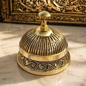 Peut inclure: Une cloche de bureau en laiton poli avec un d&ocirc;me arrondi pr&eacute;sentant des rainures verticales. La base de la cloche est orn&eacute;e d'un motif de vagues en relief. Un petit bouton circulaire lisse se trouve au sommet pour l'activation manuelle.