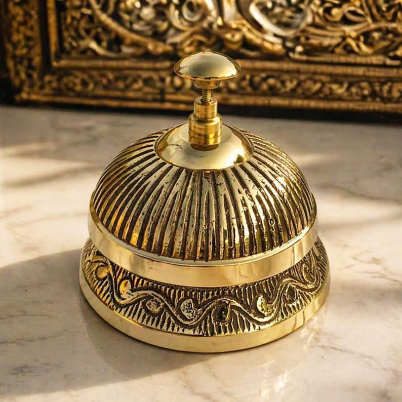 Peut inclure: Une cloche de bureau en laiton poli avec un d&ocirc;me arrondi pr&eacute;sentant des rainures verticales. La base de la cloche est orn&eacute;e d'un motif de vagues en relief. Un petit bouton circulaire lisse se trouve au sommet pour l'activation manuelle.
