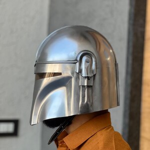 May include: A silver helmet with a visor and a T-shaped opening. The helmet has a rounded top and a chin guard. The helmet is made of a shiny, metallic material, possibly for cosplay or display. The design is reminiscent of a sci-fi character.