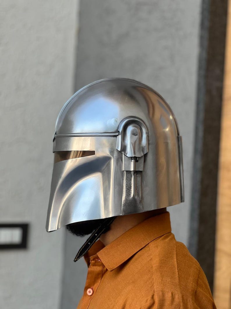 May include: A silver helmet with a visor and a T-shaped opening. The helmet has a rounded top and a chin guard. The helmet is made of a shiny, metallic material, possibly for cosplay or display. The design is reminiscent of a sci-fi character.