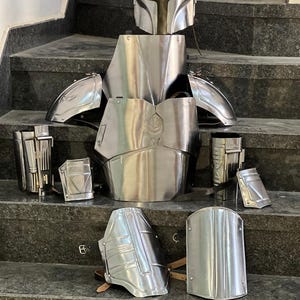 May include: A complete set of polished metal armor, including a helmet, chest plate, shoulder pads, and various other protective pieces. The armor is displayed on a set of stairs, showcasing its intricate design and craftsmanship. The armor is a silver color.