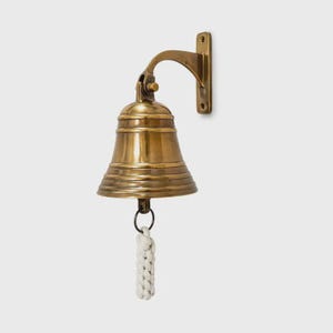 May include: A polished brass wall-mounted bell featuring a flared rim and a curved mounting bracket. A white braided rope lanyard is attached to the clapper, hanging vertically from the centre of the bell, which has a smooth, metallic finish.