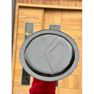 May include: A gray and black Spartan shield with a V-shaped design. The shield is round with a black rim and a gray center. The shield has a V-shaped design in the center and is held up against a wooden background.
