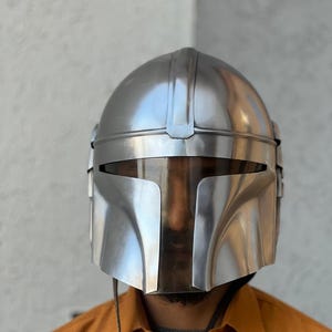 May include: A shiny, silver helmet with a T-shaped visor, evoking a futuristic warrior aesthetic. The helmet features a smooth, reflective finish and a design with a rounded crown and sharp angles. A dark cord hangs from the bottom.