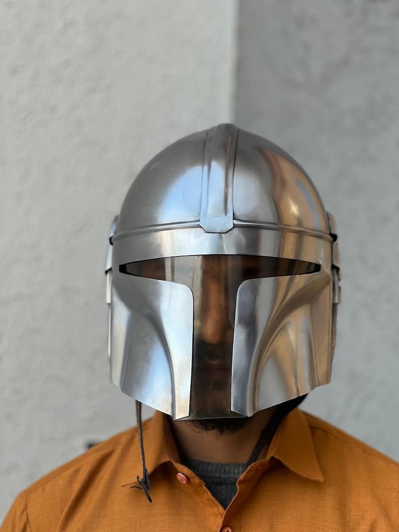 May include: A shiny, silver helmet with a T-shaped visor, evoking a futuristic warrior aesthetic. The helmet features a smooth, reflective finish and a design with a rounded crown and sharp angles. A dark cord hangs from the bottom.
