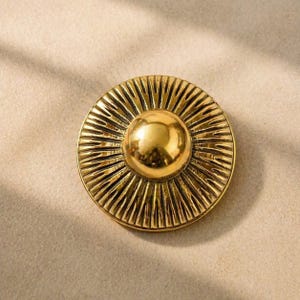 Peut inclure: Un bouton circulaire dor&eacute; dot&eacute; d'un d&ocirc;me central lisse et sur&eacute;lev&eacute;, entour&eacute; d'un motif textur&eacute; en rayons de soleil. La surface m&eacute;tallique pr&eacute;sente une finition polie avec des rainures sombres en creux entre les rayons.