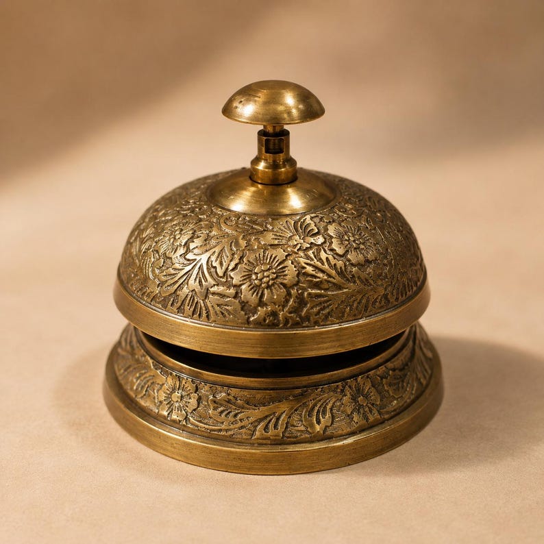Peut inclure: Une sonnette de bureau en laiton avec un motif floral en relief complexe sur le d&ocirc;me et la base. La sonnette poss&egrave;de un bouton-poussoir arrondi sur le dessus et une finition m&eacute;tallique aux tons brun dor&eacute;, orn&eacute;e de gravures de fleurs et feuilles.