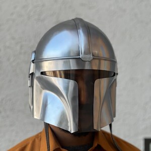 May include: A silver Mandalorian helmet with a T-shaped visor. The helmet has a rounded top and angular features, with a smooth, metallic finish. The design includes vertical lines and a curved lower section.