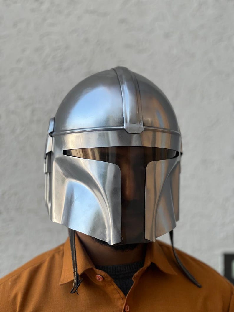May include: A silver Mandalorian helmet with a T-shaped visor. The helmet has a rounded top and angular features, with a smooth, metallic finish. The design includes vertical lines and a curved lower section.