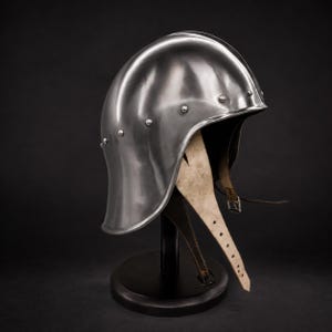 Pode incluir: Um capacete de aço polido do tipo chapéu de ferro com uma cúpula arredondada com sulcos verticais e uma aba larga. O capacete é adornado com uma série de pequenos rebites metálicos ao longo da borda inferior e inclui tiras de queixo de couro.