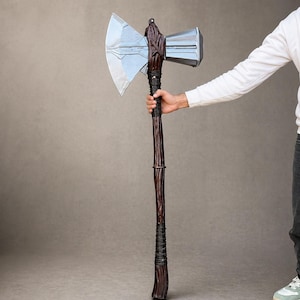 Pode incluir: Um machado de batalha de cabeça dupla com uma lâmina de prata metálica de um lado e uma cabeça de martelo retangular do outro. O cabo longo tem um acabamento de madeira texturizada em castanho escuro com detalhes pretos enrolados.
