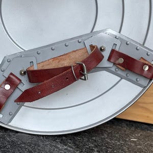 K&ouml;nnte beinhalten: Ein silberfarbener Schild mit konzentrischen Kreisen. Der Schild hat einen grauen Metallarmschutz mit Nieten und wird durch drei weinrote Lederriemen mit silbernen Schnallen und Nieten befestigt. Geeignet f&uuml;r historische Nachstellungen oder Kost&uuml;me.