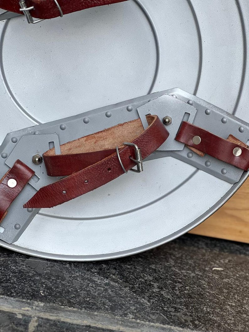 K&ouml;nnte beinhalten: Ein silberfarbener Schild mit konzentrischen Kreisen. Der Schild hat einen grauen Metallarmschutz mit Nieten und wird durch drei weinrote Lederriemen mit silbernen Schnallen und Nieten befestigt. Geeignet f&uuml;r historische Nachstellungen oder Kost&uuml;me.
