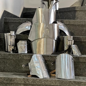 May include: A complete set of shiny, silver-colored armor, including a helmet, chest plate, shoulder pads, and various other protective pieces. The armor is displayed on a set of stairs, showcasing its intricate design and metallic finish.