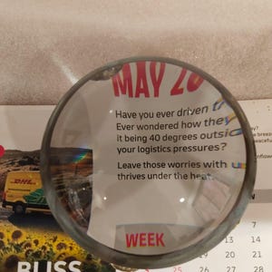 May include: A magnifying glass with a dark metal rim is positioned over a calendar page. The calendar shows the date "MAY 20" in red, along with the word "WEEK". The image also includes text about logistics.