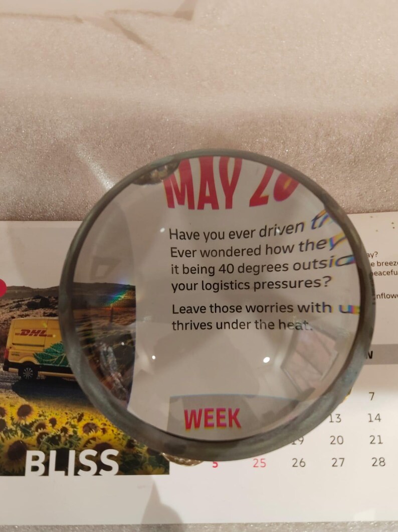 May include: A magnifying glass with a dark metal rim is positioned over a calendar page. The calendar shows the date "MAY 20" in red, along with the word "WEEK". The image also includes text about logistics.
