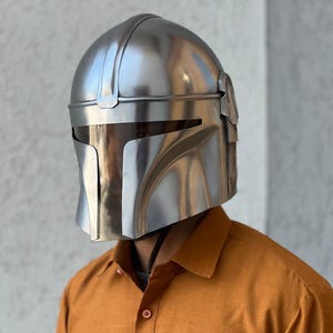 May include: A polished silver Mandalorian helmet. The helmet features a distinctive T-shaped visor and angular design. The helmet is made of metal and reflects the light. The overall design is sleek and futuristic.