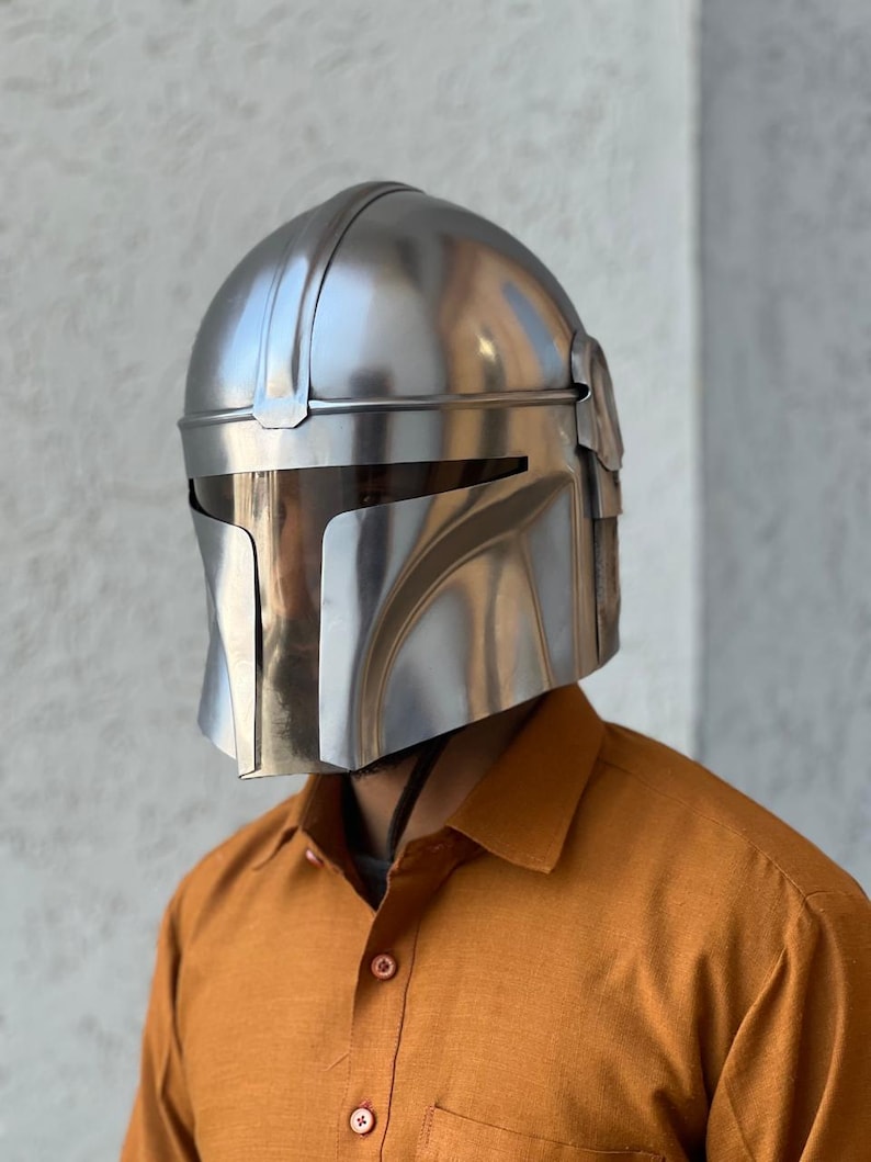 May include: A polished silver Mandalorian helmet. The helmet features a distinctive T-shaped visor and angular design. The helmet is made of metal and reflects the light. The overall design is sleek and futuristic.