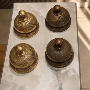 Peut inclure: Quatre sonnettes de comptoir en laiton dispos&eacute;es en carr&eacute;. Deux sonnettes pr&eacute;sentent des d&eacute;tails nervur&eacute;s verticaux, tandis que les deux autres arborent des gravures florales complexes. Chaque sonnette poss&egrave;de une base circulaire.