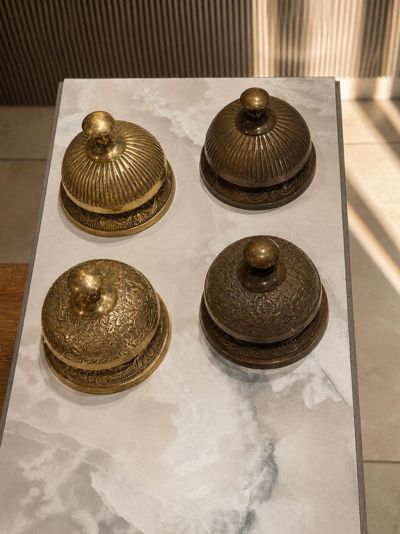 Peut inclure: Quatre sonnettes de comptoir en laiton dispos&eacute;es en carr&eacute;. Deux sonnettes pr&eacute;sentent des d&eacute;tails nervur&eacute;s verticaux, tandis que les deux autres arborent des gravures florales complexes. Chaque sonnette poss&egrave;de une base circulaire.